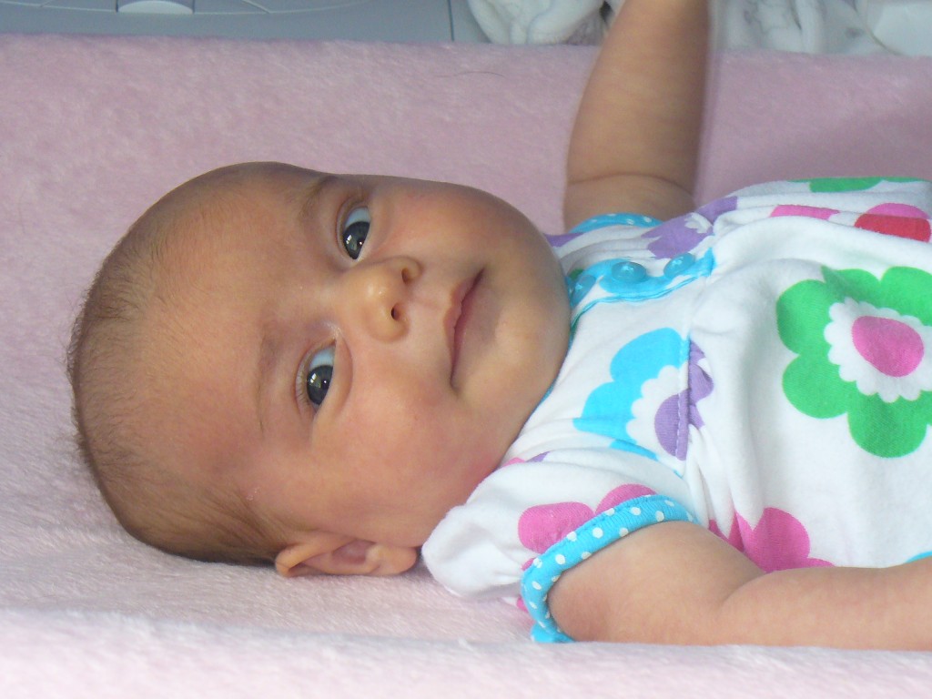 What is it like to be a new SAHM? A baby wearing a white outfit with colorful flower patterns lies on a light pink blanket. The baby is looking slightly to the side with a calm expression. One arm is resting by their side, and the other is slightly raised. This serene moment captures the quiet joys stay-at-home moms cherish in their daily routines.