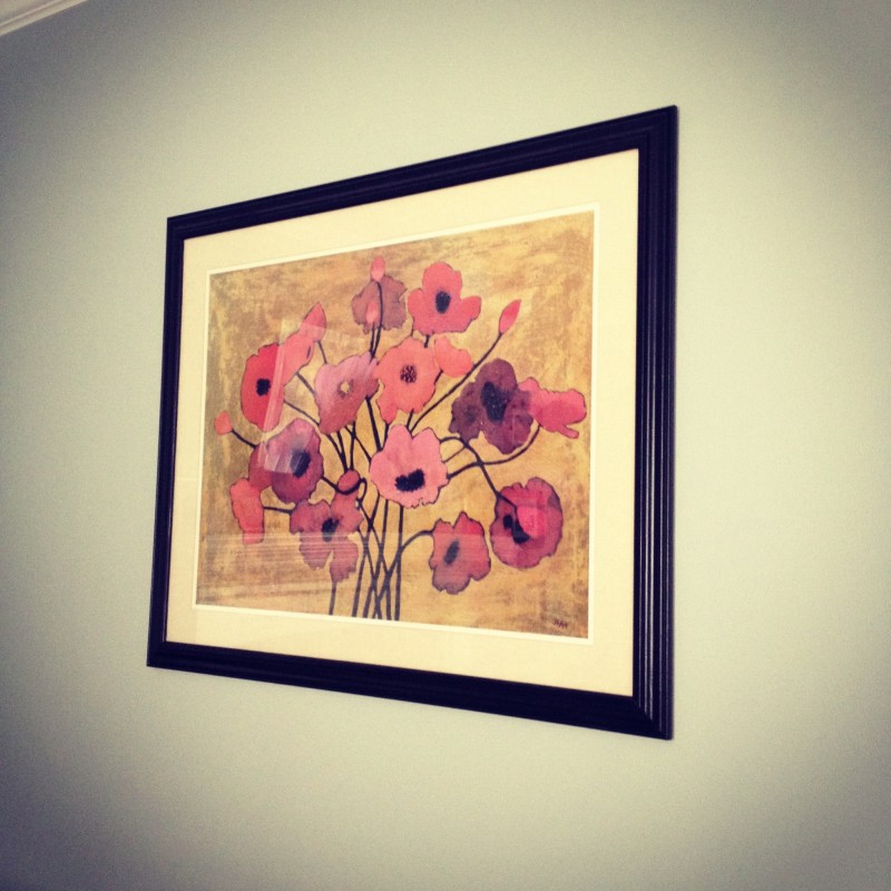 A framed painting of pink and purple flowers on a tan background hangs on a light green wall. The artwork, often found among the best deals at garage sales, is bordered by a black frame with a white mat. The abstract flowers have long stems and various shapes and shades.