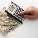 A person's hand using a black and white calculator placed on top of several U.S. dollar bills spread out on a light-colored surface. The scene implies financial calculations or budgeting for monthly household expenses.