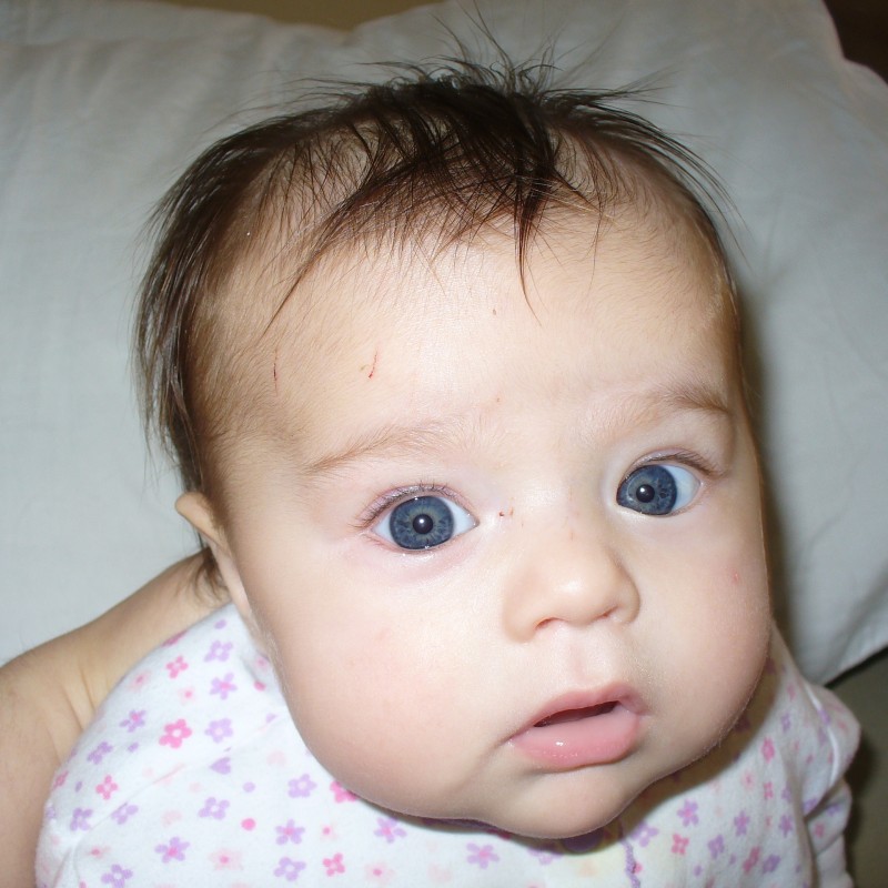 A baby with blue eyes and slightly messy hair is looking at the camera with a curious expression.