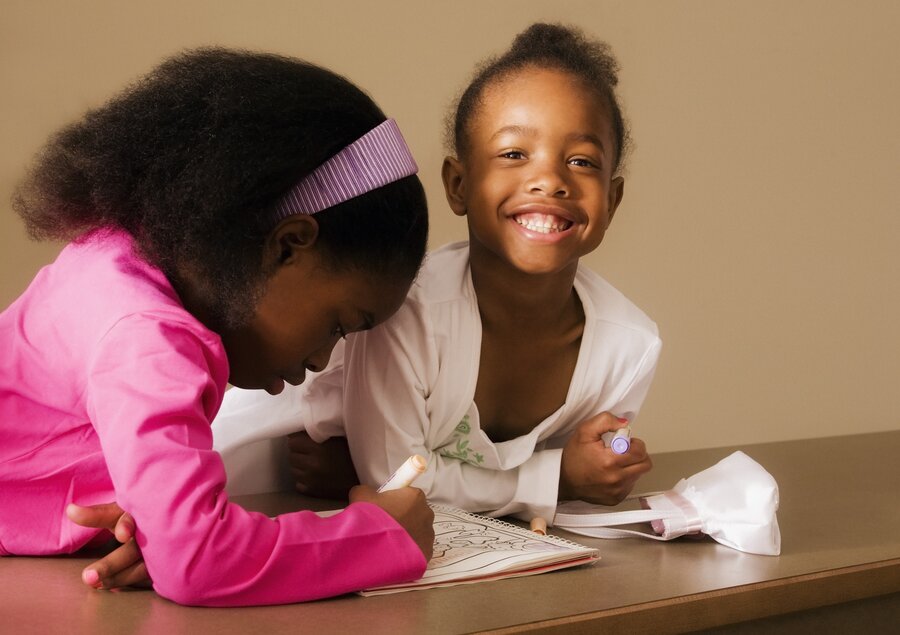 Two young Black little girl in pink shirts coloring on a sheet of paper having fun.