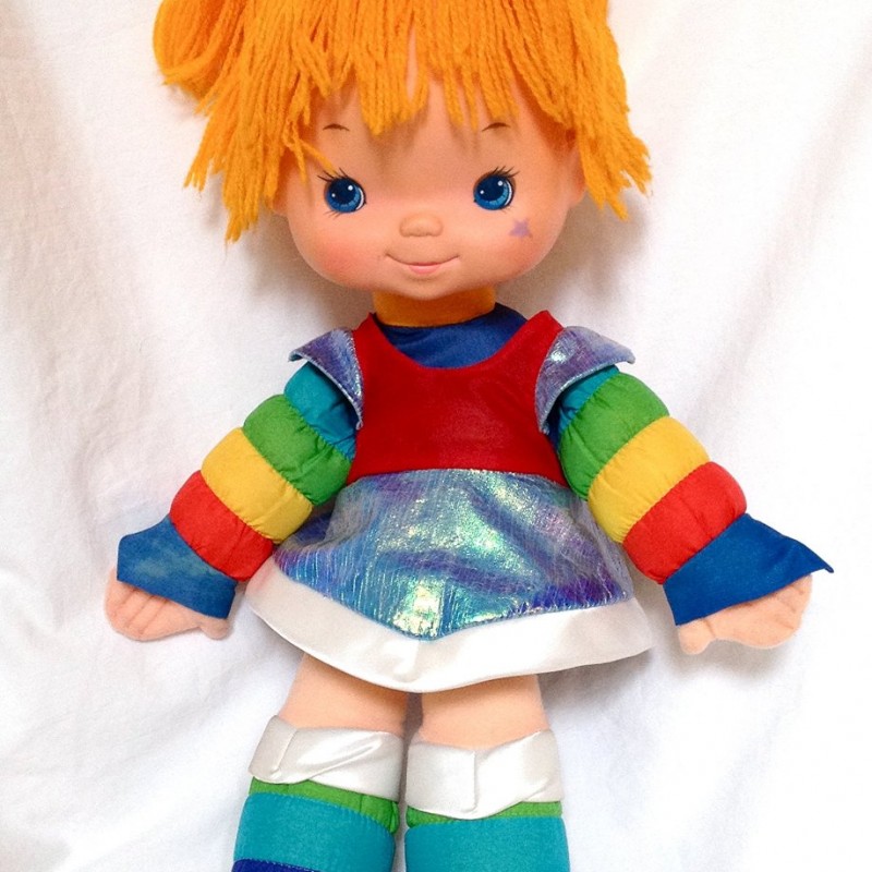 An adorable, vintage 1980s doll dressed in a vibrant, multicolored outfit stands against a white background. The doll has bright orange yarn hair tied with a blue ribbon, blue eyes, and a cheerful expression. The outfit includes rainbow-striped sleeves and leggings.