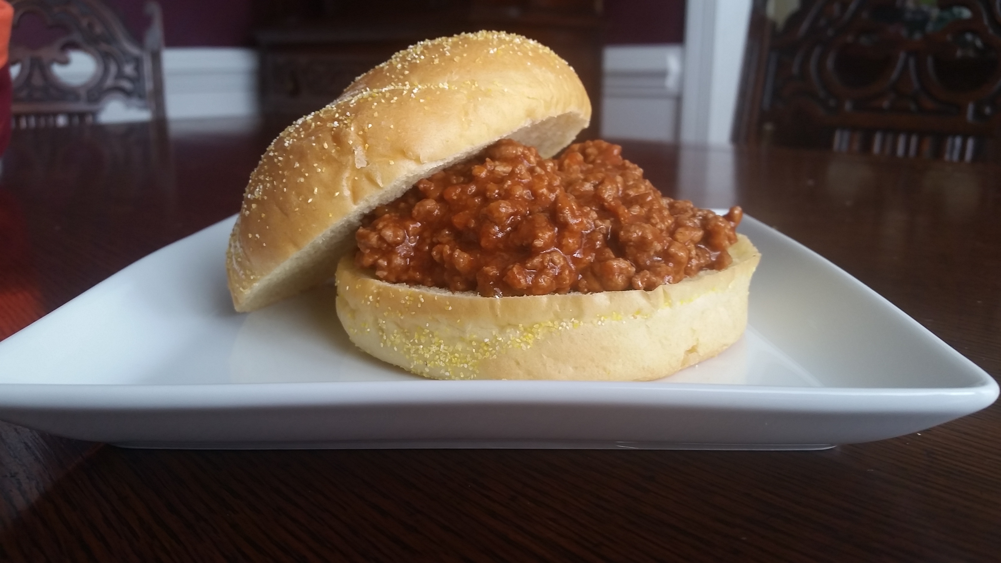 A sandwich with a generous filling of seasoned ground meat is served on a white plate. The bun has a slightly yellow, crumbly texture on the outside, and the sandwich sits in focus on a dark wooden table with ornate, blurred chairs in the background.