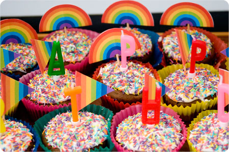 Colorful cupcakes adorned with rainbow sprinkles and rainbow flags. Each cupcake has a letter-shaped candle, collectively spelling out "HAPPY BIRTHDAY." The cupcakes are placed in vibrant, multicolored wrappers, and free rainbow printables enhance the festive, cheerful atmosphere.