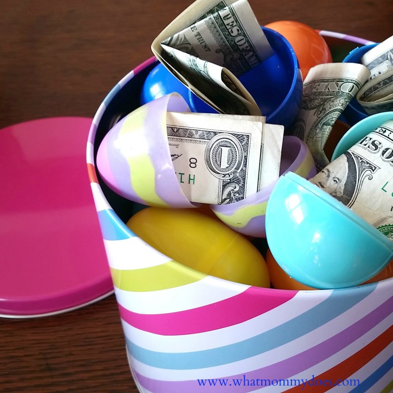 A colorful, striped container with a pink lid is filled with plastic eggs of various colors, some open to reveal U.S. dollar bills inside—a perfect money gift idea. The container is placed on a wooden surface.