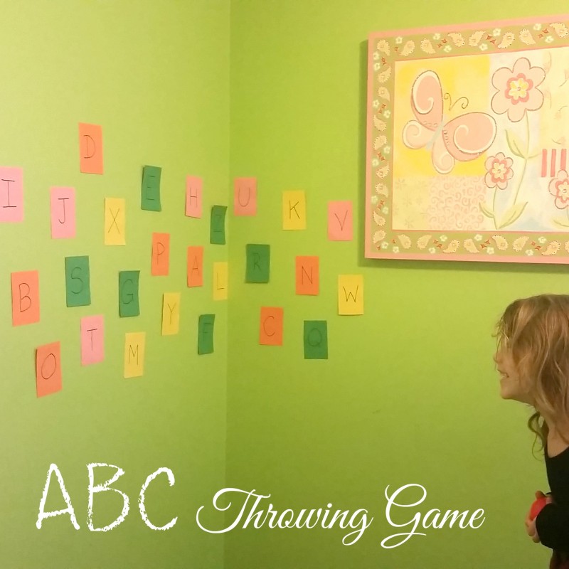A young child with long hair stands in front of a green wall, aiming at colorful paper squares with letters from the alphabet written on them. A floral and butterfly-themed picture hangs on the wall, and "ABC Throwing Game" is prominently displayed at the bottom.