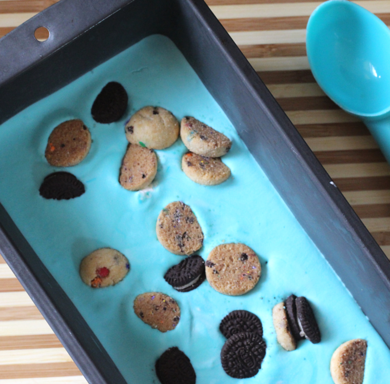 A rectangular baking tin filled with cookie monster ice cream is shown. The blue ice cream is topped with assorted mini cookies and small Oreo cookies. A matching blue scoop rests nearby on a wooden, striped surface.
