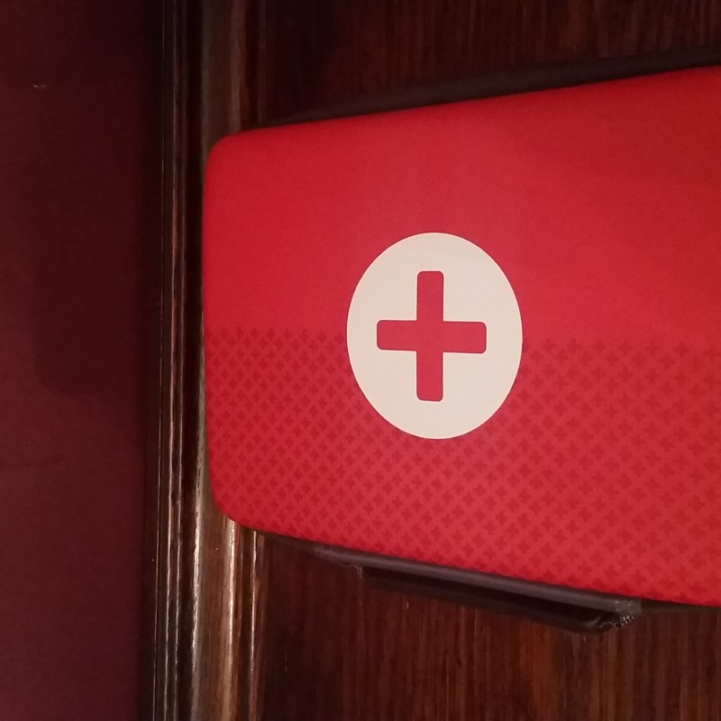 A red first aid kit with a white cross emblem on the cover. The kit, which doubles as an ideal emergency stash, has a dark handle on the side and is placed on a wooden surface with a maroon wall in the background.