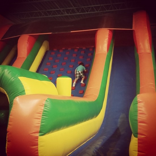 A child wearing a green shirt and plaid shorts climbs a colorful inflatable obstacle course with red, green, yellow, and blue sections. The child is scaling a wall with red grips in an indoor play area, capturing a typical adventurous moment in the life of a stay-at-home-mom blogger.