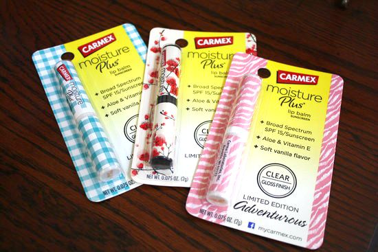 Three stylish Carmex Moisture Plus lip balm packages are displayed. Each package boasts a unique design: blue gingham, red floral, and pink zigzag patterns. The packaging highlights features like SPF 15, aloe, vitamin E, and a clear gloss finish with various scent choices.