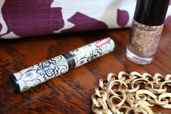 A close-up of a table featuring a white eyeliner pen with a black floral design, a bottle of glittery nail polish, a chunky gold bracelet, and stylish lip balm next to purple and white patterned fabric in the background.