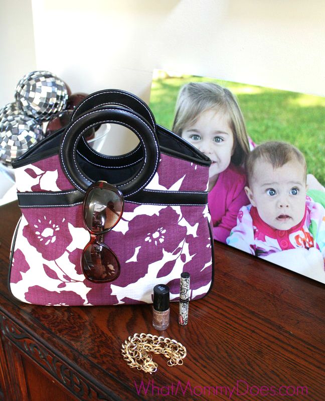 A handbag with circular handles and a purple and white floral pattern is placed on a wooden table. The bag, perfect for things to sell quickly, has sunglasses hooked onto the handle. Nearby, there are makeup items, a bracelet, and a framed photo of two children.