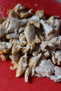 Chopped cooked chicken pieces on a red cutting board. The chicken appears to be lightly seasoned and shredded into small chunks, ideal for crockpot buffalo chicken dip or any meal preparation.