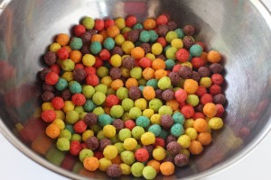 A stainless steel bowl filled with a mix of colorful, round cereal pieces sits on a white surface. The cereal pieces are red, orange, yellow, green, blue, and purple, creating a vibrant, multicolored display reminiscent of Halloween treats on a spooky night.