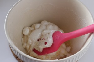 A large white bowl contains a mixture of melted marshmallow and butter or another ingredient, setting the stage for an Eye Popping Cake. A red spatula is partially embedded in the mixture, showing some of the sticky, gooey texture. The bowl is placed on a cork mat.