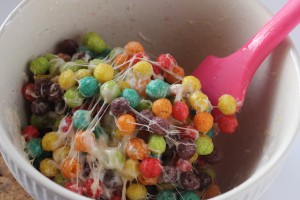 A white bowl filled with colorful cereal balls mixed with melted marshmallows looks like a Halloween treat. A pink spatula is sticking out, used for mixing the sticky, spooky concoction. The cereal balls are yellow, orange, red, green, and purple.