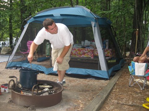 A man stirs a large pot over a fire pit at a campsite. Behind him is a blue and white screened tent with a table inside, covered with a colorful cloth. Trees surround the campsite, and camping chairs are scattered around the area.