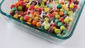 A close-up view of a glass dish filled with colorful, round cereal pieces coated in melted marshmallow strings. The mixture includes vibrant red, green, yellow, orange, and purple cereal pieces, giving a sweet and playful appearance reminiscent of a spooky Halloween bundt cake.