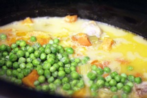 A close-up of a slow cooker meal in progress, showing a mixture of ingredients including fresh green peas, chopped orange carrots, celery pieces, and chunks of meat in a creamy, yellowish broth resembling crockpot chicken pot pie.
