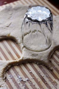 A glass is being used to cut circles out of rolled-out dough on a wooden surface, for what looks like a crockpot chicken pot pie. The dough has a few circles already cut out, and the glass is placed upside down, pressing into the dough. The scene is lit by natural light.