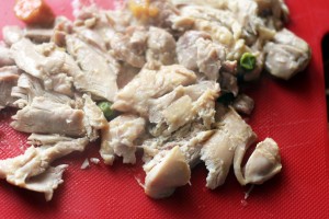 A close-up of chopped cooked chicken pieces on a red cutting board, reminiscent of crockpot chicken pot pie. The chunks of white and light brown meat are scattered, with some small bits of vegetables like green peas and carrots visible among them.