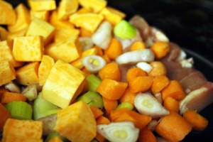 A close-up image of chopped vegetables in a cooking dish, resembling ingredients for a crockpot chicken pot pie. The vegetables include large cubes of yellow sweet potatoes, small chunks of orange carrots, slices of green celery, and pieces of white onions. The dish appears to be ready for cooking.