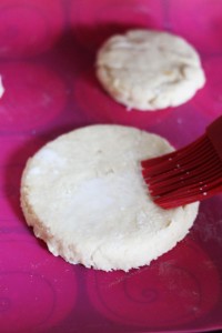 A close-up of dough rounds on a pink surface. A red silicone pastry brush is applying an egg wash or similar liquid to one of the dough rounds, perhaps preparing them for a crockpot chicken pot pie. The dough appears to be for biscuits or scones.