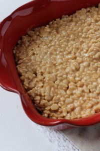 A close-up of a red baking dish filled with homemade rice cereal treats. The treats appear gooey and slightly golden, with visible puffed rice cereal bits, indicating they were freshly made and are ready to be enjoyed.