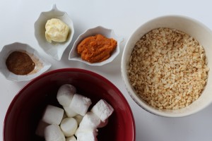 Several ingredients are laid out for making a sweet treat. There is a bowl of rice cereal, a red bowl filled with large marshmallows, and small dishes containing butter, pumpkin puree, and pumpkin pie spice.