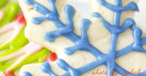 A close-up view of a snowflake-shaped sugar cookie decorated with blue icing, perfect for an easy Christmas cookie exchange. Other cookies with green and pink icing are visible in the background. The photo credit "photo by: iyoubal" is written in red text at the bottom right corner.