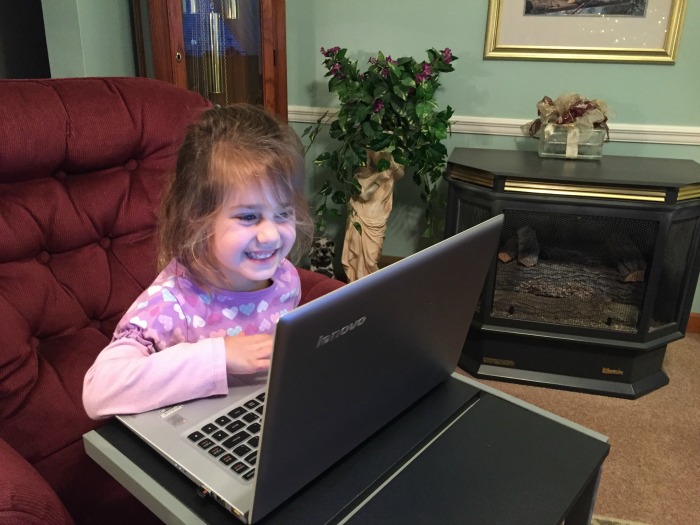 A young girl with long brown hair, wearing a pink and purple top, is sitting on a maroon chair, smiling and looking at a laptop screen in front of her as she connects with family. The room includes a fireplace, a plant, and a framed picture on the wall.