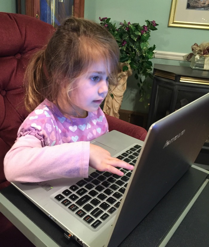 A young girl with light brown hair in a ponytail is focused on a laptop in front of her, perhaps trying to connect with family. She is wearing a pink and purple shirt with heart patterns, seated on a maroon chair in a room with a green plant and a picture frame in the background.