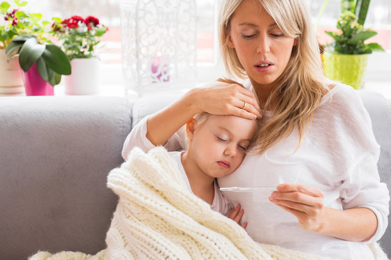 A woman is sitting on a couch, gently holding a thermometer and placing her hand on the forehead of a sleeping child wrapped in a white blanket. The background features potted plants and a large window letting in natural light.