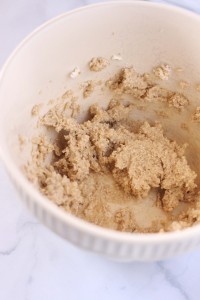A white bowl with ridged edges contains a grainy beige mixture, likely from an easy Christmas cookie recipe, sitting on a white marble surface. The mixture appears to be partially mixed, with some clumps and a slightly uneven texture.