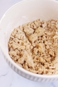 A ceramic bowl filled with crumbly dough mixture, indicating the initial stages of an easy Christmas cookie recipe. The dough appears coarse and unevenly mixed with visible chunks of butter and flour. The bowl is placed on a white, marble-like surface.