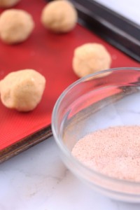 A close-up of cookie dough balls placed on a red silicone baking mat on a baking sheet. In the foreground, there is a small glass bowl containing a cinnamon-sugar mixture. The background is slightly blurred, emphasizing the cookie preparation process—a perfect scene for an easy Christmas cookie recipe.