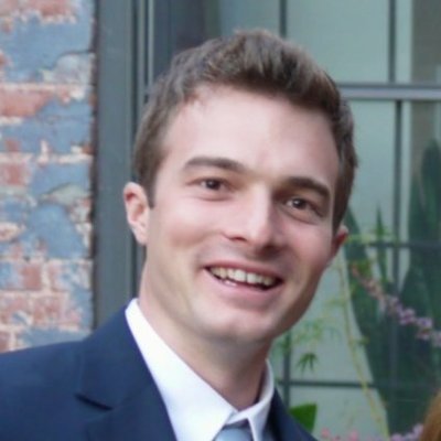 A man with short brown hair, wearing a navy blue suit, white dress shirt, and light blue tie, smiles at the camera. He is standing in front of a window with a brick wall on the side and some greenery visible in the background—an example of effective SEO strategies yielding 17K to 400K+ page views over 10 months.