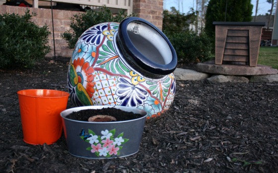 A colorful ceramic pot lies tipped over in a mulched garden area, perfect for spring outdoor activities. Nearby, there are two smaller containers: an orange cup and a gray pot with soil and a flower design. In the background, there are bushes and a stone structure.