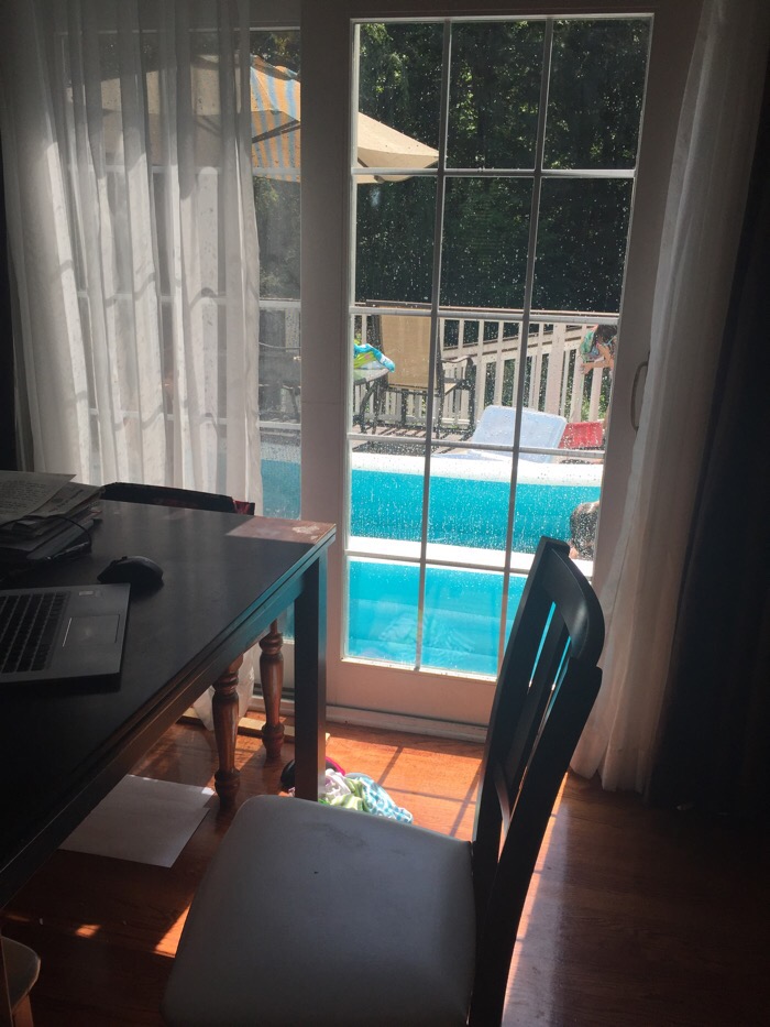 A workspace setup inside a room with a table, chair, and laptop is pictured near a glass door. Outside, there's a swimming pool and deck chairs, partially visible through the glass door. Sunlight filters in, creating a cozy indoor atmosphere as you contemplate the perfect moment to pick a blog name.
