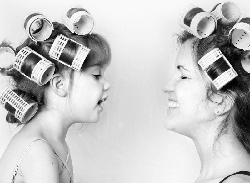 A young girl and woman, both with hair curlers, are facing each other. The girl appears to be talking, possibly recounting a tale of why kids misbehave, while the woman is smiling, showing a joyful interaction. The black-and-white image emphasizes their expressions and the curlers in their hair.