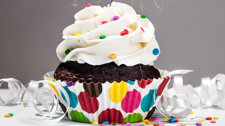 A chocolate cupcake with white frosting, adorned with colorful sprinkles, sits in a polka-dot paper liner. White ribbon adornments and scattered sprinkles surround the cupcake, creating a festive setting reminiscent of birthday freebies. The background is a plain grey.