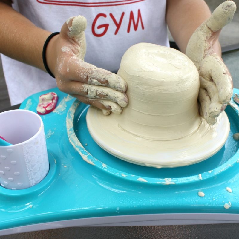 A person shaping a clay bowl on a blue pottery wheel. Their hands are covered in clay, and they are wearing a white shirt with the word "GYM" visible. The setup looks perfect for kids activities, with a cup holding a brush beside the wheel—a delightful introduction to hobbies for kids.