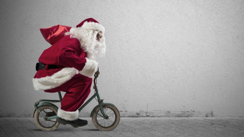 A person dressed as Santa Claus rides a small, worn bicycle against a plain, textured wall. The kids would believe it's truly Santa, crouched low and creating a humorous contrast between his large frame and the tiny bike.
