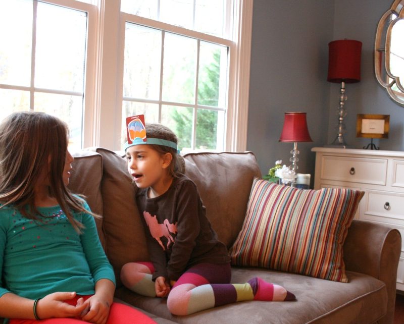 Two young girls sit on a couch in a cozy living room with large windows. One girl wears a headband with a card on her forehead and gestures animatedly, making it an easy family game night idea. The other girl listens attentively. The room has a striped cushion, red lamp, white dresser, and wall mirror.