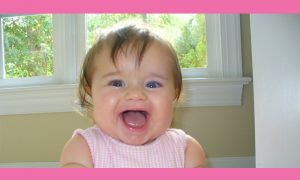 A cheerful baby in a pink sleeveless outfit is laughing, with her mouth open wide and eyes bright, as if she's just heard some fun money management tips. She is sitting in front of a window with greenery visible outside. The photo is framed by a pink border.