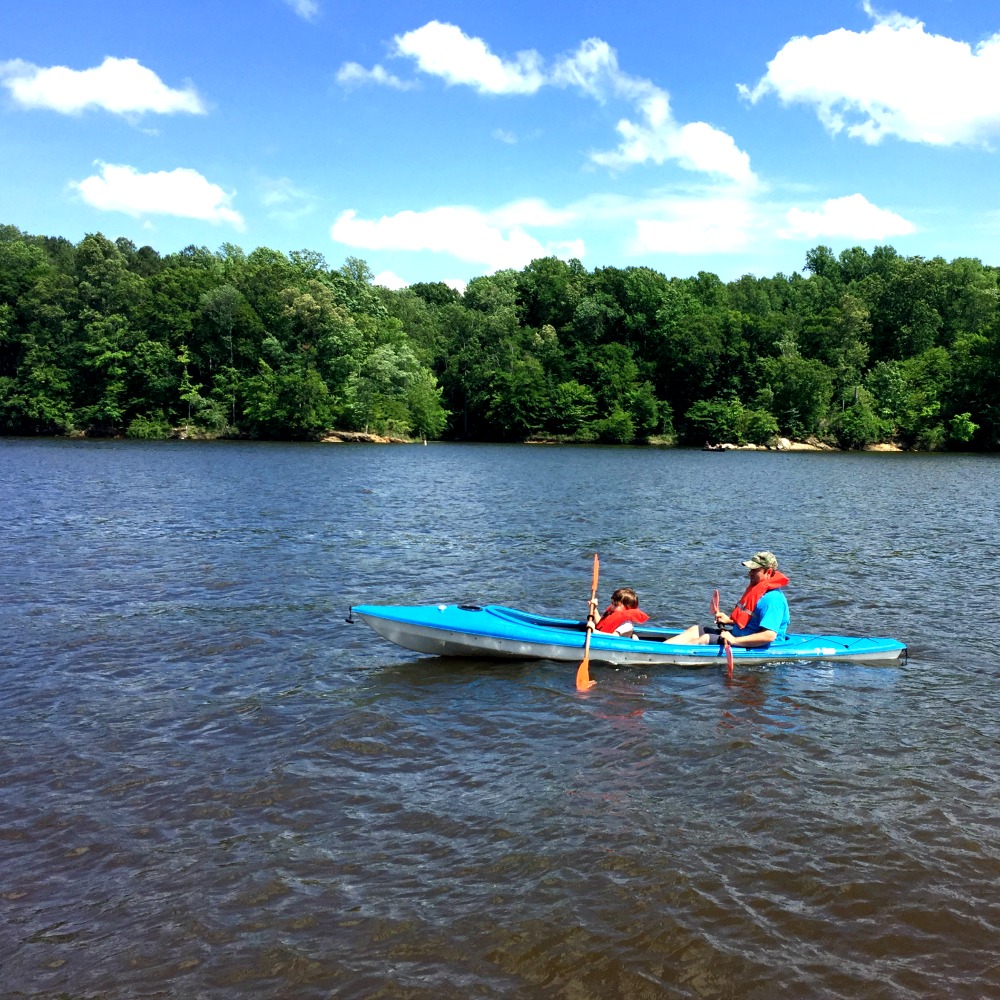 Two people wearing life vests paddle in a blue kayak on a calm lake surrounded by dense, green trees under a bright blue sky with scattered clouds. Unplugging from daily routines, they enjoy their weekend adventure as the person in the front uses a double-bladed paddle, moving the kayak away from the camera.