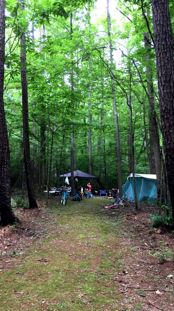 A campsite nestled in a forest clearing features a blue tent on the right and a canopy with a picnic table beneath it in the background. Bicycles and camping gear are scattered around, surrounded by tall trees with lush green foliage—perfect for unplugging on a weekend adventure.