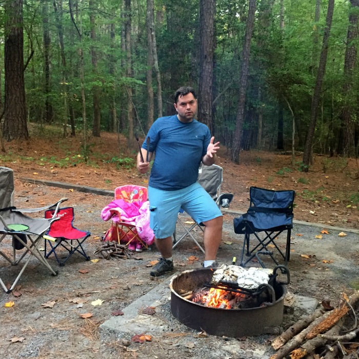A man in a blue shirt and shorts stands next to a campfire pit surrounded by chairs in a forested area. He is posing with a playful expression, holding a grilling fork and what appears to be a plate or pan, perhaps preparing his favorite campfire nachos recipe. There is a blanket and a bundle of sticks nearby.