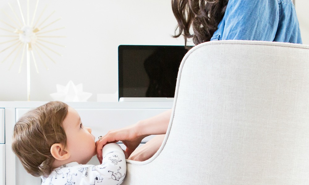A baby, dressed in a white outfit with animal prints, leans against a white chair and holds the hand of a seated woman with long brown hair, partially visible, wearing a blue shirt. In the background, where she finds moments of solace from her blogging overwhelm, a laptop and several decorative items are on a desk.