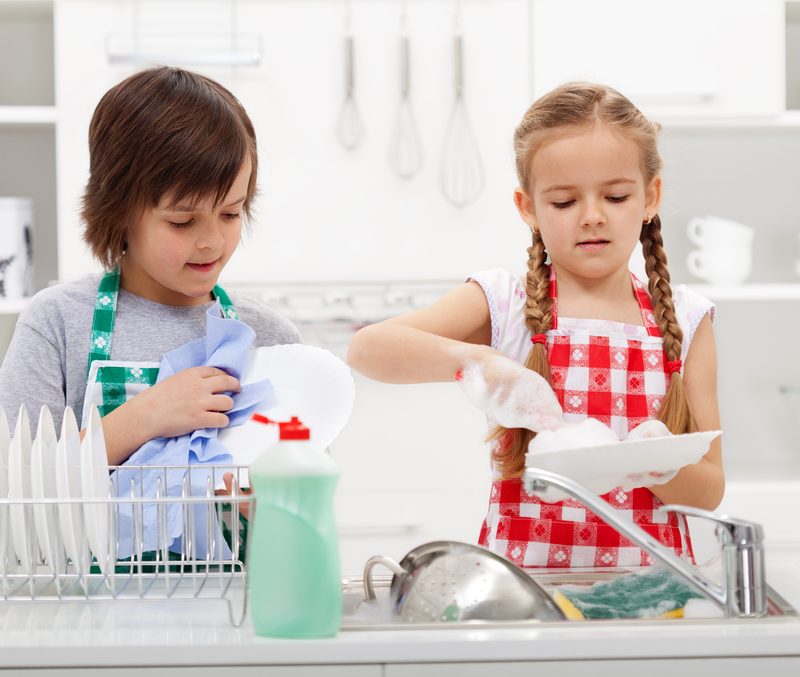 Two children are washing dishes in a bright kitchen. The boy on the left, wearing a green apron, dries a plate with a blue cloth, while the girl on the right, wearing a red checkered apron, washes a plate with a sponge over the sink. A blank printable weekly chore chart is taped to the fridge beside them.